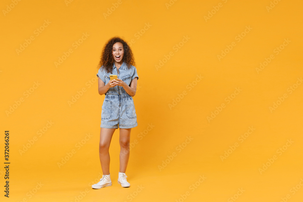 Full length portrait of excited young african american woman girl in denim clothes isolated on yellow wall background in studio. People lifestyle concept. Using mobile cell phone, typing sms message. Full length portrait of excited young african american woman girl in denim clothes isolated on yellow wall background in studio. People lifestyle concept. Using mobile cell phone, typing sms message.