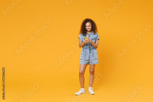 Full length portrait of smiling young african american woman girl in denim clothes isolated on yellow wall background in studio. People lifestyle concept. Using mobile cell phone, typing sms message.