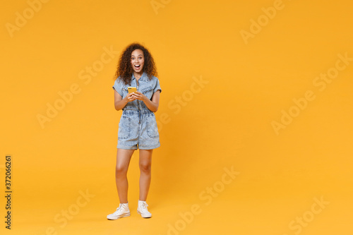 Full length portrait of excited young african american woman girl in denim clothes isolated on yellow wall background in studio. People lifestyle concept. Using mobile cell phone, typing sms message.
