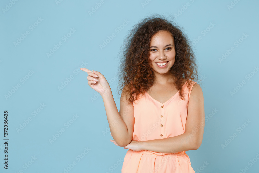 Smiling young african american girl in pastel summer clothes posing isolated on blue background studio portrait. People emotions lifestyle concept. Mock up copy space. Pointing index finger aside. Smiling young african american girl in pastel summer clothes posing isolated on blue background studio portrait. People emotions lifestyle concept. Mock up copy space. Pointing index finger aside.