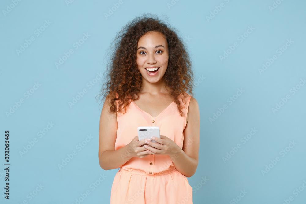Excited young african american girl in pastel summer clothes isolated on blue wall background studio portrait. People lifestyle concept. Mock up copy space. Using mobile cell phone typing sms message. Excited young african american girl in pastel summer clothes isolated on blue wall background studio portrait. People lifestyle concept. Mock up copy space. Using mobile cell phone typing sms message.