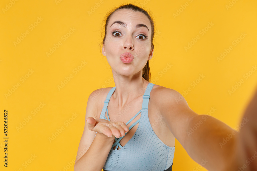 Close up of pretty fitness sporty woman in sportswear working out isolated on yellow background. Workout sport motivation lifestyle concept. Doing selfie shot on mobile phone blowing sending air kiss.