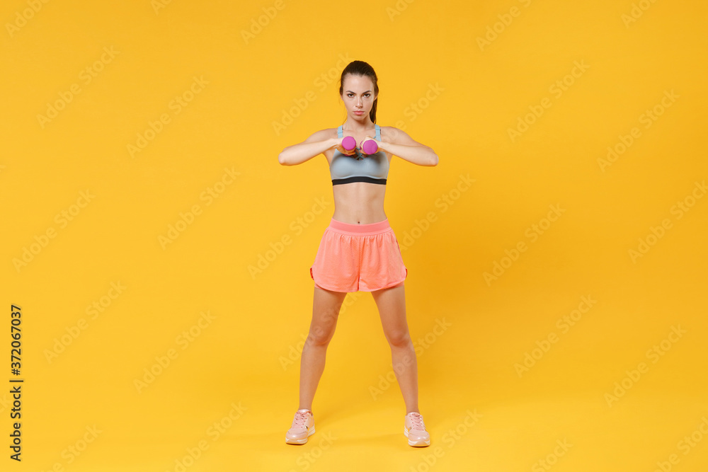 Full length portrait of serious young fitness woman girl in sportswear working out isolated on yellow wall background studio. Workout sport motivation lifestyle concept. Doing exercise with dumbbell. Full length portrait of serious young fitness woman girl in sportswear working out isolated on yellow wall background studio. Workout sport motivation lifestyle concept. Doing exercise with dumbbell.