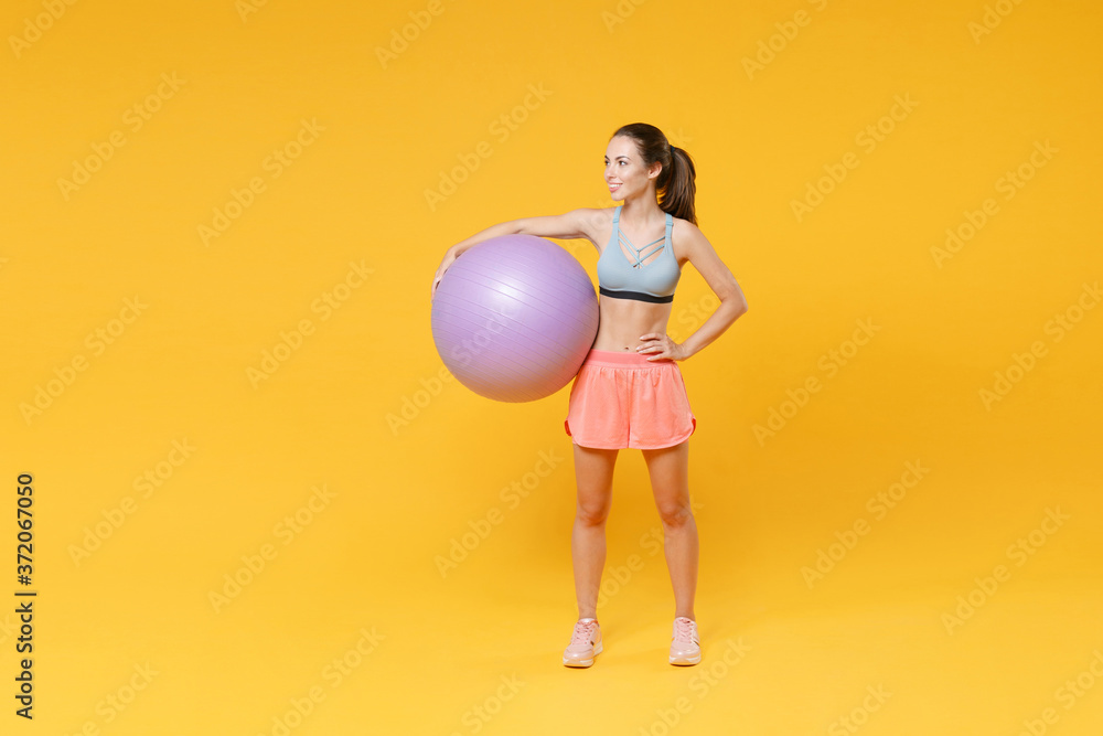 Full length portrait of smiling young fitness woman girl in sportswear working out isolated on yellow wall background studio. Workout sport motivation lifestyle concept. Hold fitball, looking aside. Full length portrait of smiling young fitness woman girl in sportswear working out isolated on yellow wall background studio. Workout sport motivation lifestyle concept. Hold fitball, looking aside.