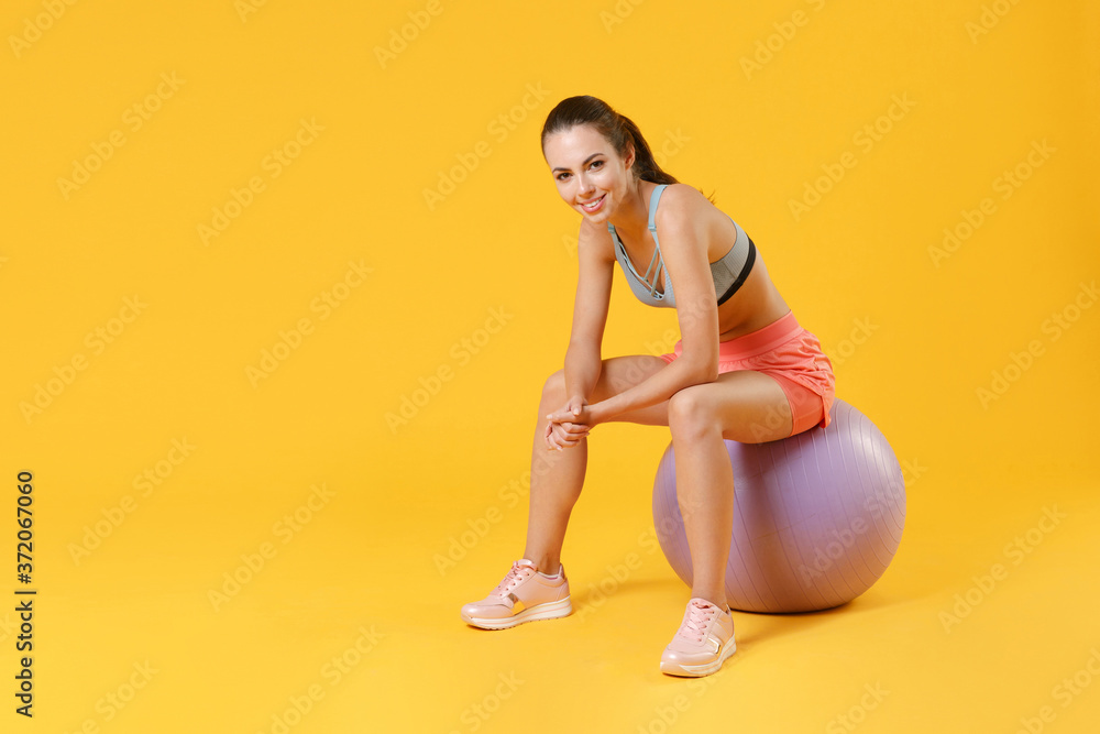 Full length portrait of smiling young fitness sporty woman girl in sportswear working out isolated on yellow background studio portrait. Workout sport motivation lifestyle concept. Sit on fitball. Full length portrait of smiling young fitness sporty woman girl in sportswear working out isolated on yellow background studio portrait. Workout sport motivation lifestyle concept. Sit on fitball.