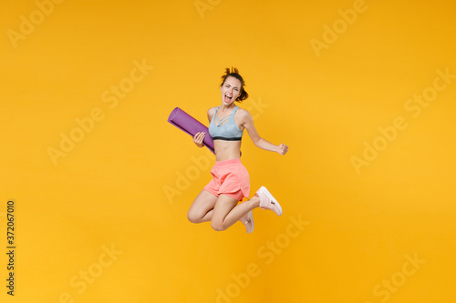 Full length portrait screaming young fitness woman in sportswear working out isolated on yellow background. Workout sport motivation lifestyle concept. Jumping, hold yoga mat, doing winner gesture.