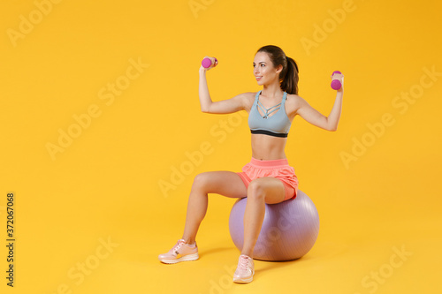 Full length portrait pretty young fitness sporty girl in sportswear working out isolated on yellow background. Workout sport motivation lifestyle concept. Sit on fitball doing exercise with dumbbell.