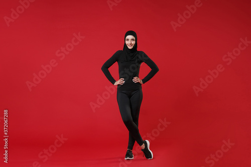 Full length portrait of smiling young arabian muslim woman in hijab black clothes posing isolated on red wall background studio. People religious lifestyle concept. Standing with arms akimbo on waist.