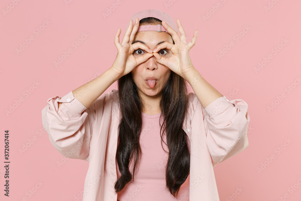 Funny young asian woman girl in casual clothes cap posing isolated on pink background. People lifestyle concept. Holding hands near eyes, imitating glasses or binoculars, making stick tongue out sign.