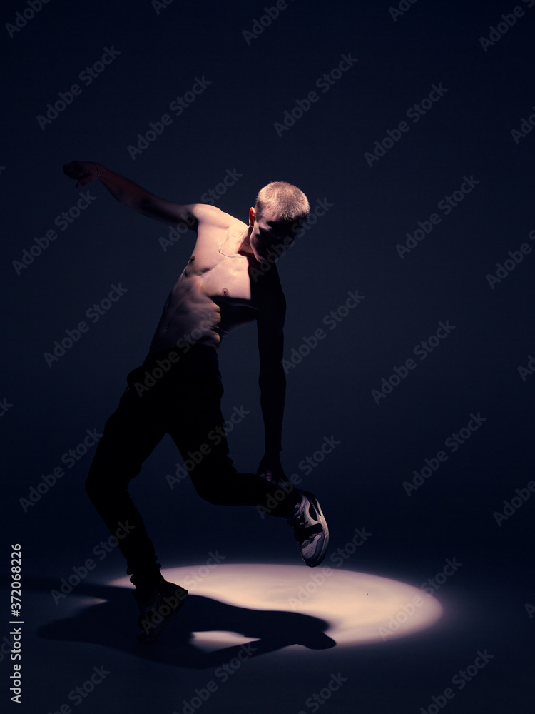 Fototapeta premium Cool young guy dancer with naked torso dancing in studio in the spotlight. Dance school poster