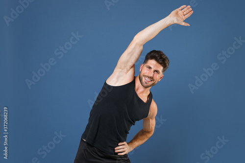 Smiling young bearded fitness sporty strong guy sportsman in black undershirt isolated on blue wall background studio portrait. Workout sport motivation lifestyle concept. Doing stretching exercising.
