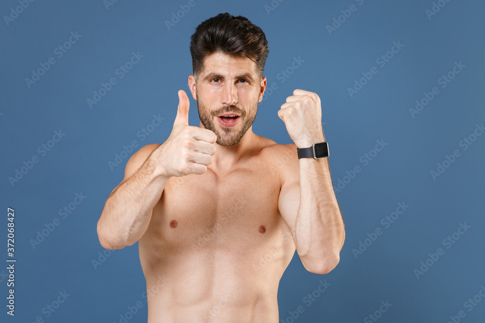 Fototapeta premium Young bearded fitness sporty strong guy bare-chested muscular sportsman posing isolated on blue background studio. Workout sport motivation lifestyle concept. Wearing smart watch, showing thumb up.