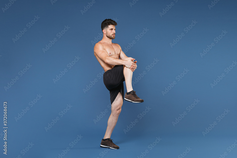 Fototapeta premium Full length portrait of young fitness sporty strong guy bare-chested muscular sportsman isolated on blue background. Workout sport motivation lifestyle concept. Doing stretching exercising for legs.