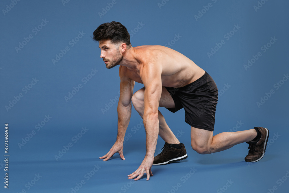 Full length portrait side view of young bearded fitness sporty strong guy bare-chested muscular sportsman isolated on blue background. Workout sport motivation lifestyle concept. Getting ready to run. Full length portrait side view of young bearded fitness sporty strong guy bare-chested muscular sportsman isolated on blue background. Workout sport motivation lifestyle concept. Getting ready to run.
