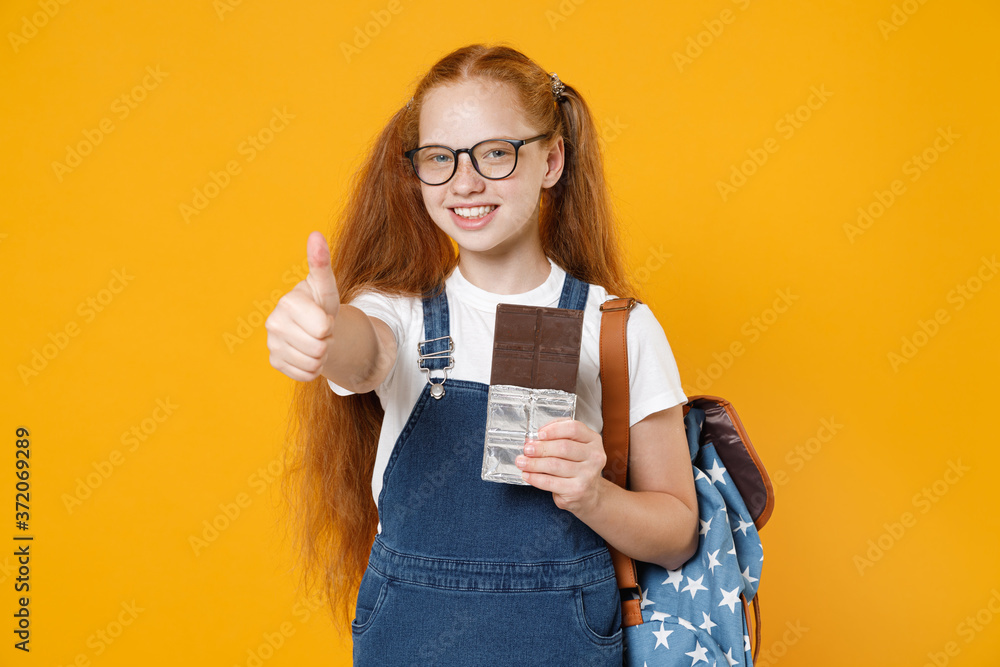Young fun redhead school teen kid girl 12-13 years old in white t-shirt blue uniform eyeglasses backpack hold eat chocolate bar isolated on yellow background children studio portrait Education concept Young fun redhead school teen kid girl 12-13 years old in white t-shirt blue uniform eyeglasses backpack hold eat chocolate bar isolated on yellow background children studio portrait Education concept