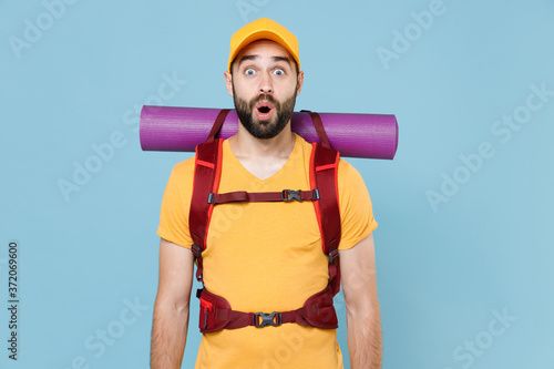 Shocked traveler young man in yellow casual t-shirt, cap with backpack isolated on blue background studio. Tourist traveling on weekend getaway. Tourism discovering hiking concept. Keeping mouth open.