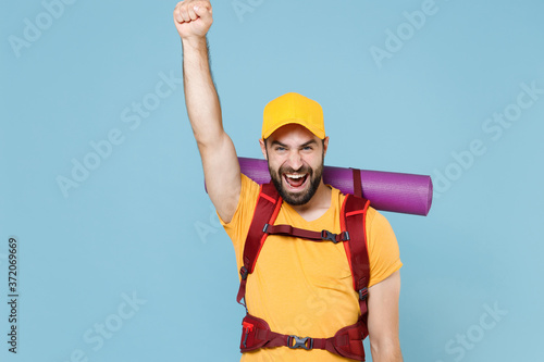 Happy traveler young man in yellow casual t-shirt cap with backpack isolated on blue background. Tourist traveling on weekend getaway. Tourism discovering hiking concept. Clenching fist like winner.