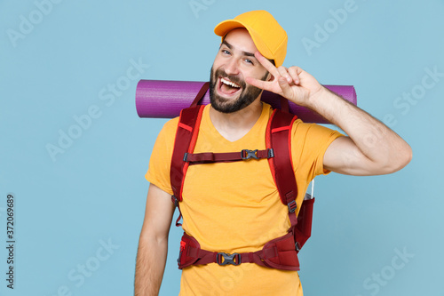 Cheerful traveler young man in yellow casual t-shirt cap with backpack isolated on blue wall background. Tourist traveling on weekend getaway. Tourism discovering hiking concept. Showing victory sign.
