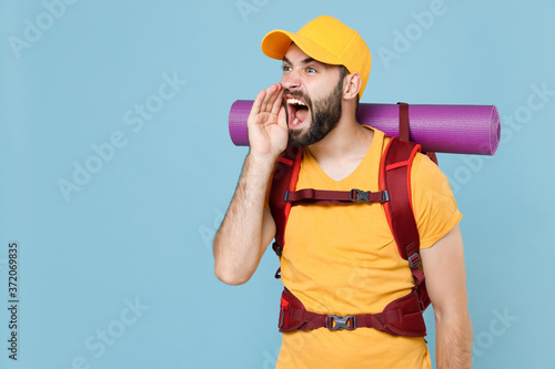 Nervous traveler young man in yellow t-shirt cap backpack isolated on blue background. Tourist traveling on weekend getaway. Tourism discovering hiking concept. Screaming with hand gesture near mouth.