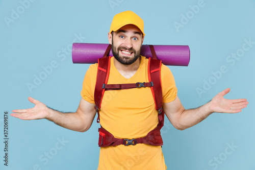 Confused traveler young man in yellow casual t-shirt cap with backpack isolated on blue background studio. Tourist traveling on weekend getaway. Tourism discovering hiking concept. Spreading hands.