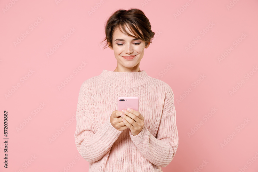Smiling young brunette woman girl wearing knitted casual sweater posing isolated on pastel pink wall background studio portrait. People lifestyle concept. Using mobile cell phone, typing sms message.