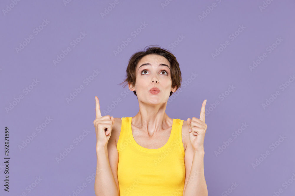 Shocked young brunette woman girl in yellow casual tank top posing isolated on pastel violet wall background studio portrait. People lifestyle concept. Mock up copy space. Pointing index fingers up. Shocked young brunette woman girl in yellow casual tank top posing isolated on pastel violet wall background studio portrait. People lifestyle concept. Mock up copy space. Pointing index fingers up.