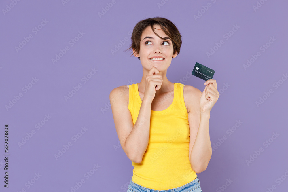 Pensive young brunette woman girl in yellow casual tank top posing isolated on pastel violet wall background studio portrait. People lifestyle concept. Hold credit bank card put hand prop up on chin. Pensive young brunette woman girl in yellow casual tank top posing isolated on pastel violet wall background studio portrait. People lifestyle concept. Hold credit bank card put hand prop up on chin.