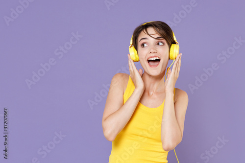 Excited young brunette woman girl wearing yellow casual tank top posing isolated on pastel violet background studio portrait. People sincere emotions lifestyle concept. Listen music with headphones. © ViDi Studio