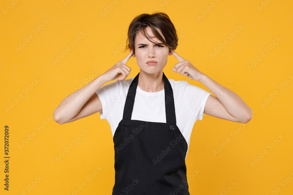 Fototapeta premium Dissatisfied young female woman 20s barista bartender barman employee in white t-shirt apron working in coffee shop covering ears with fingers isolated on yellow color wall background studio portrait.