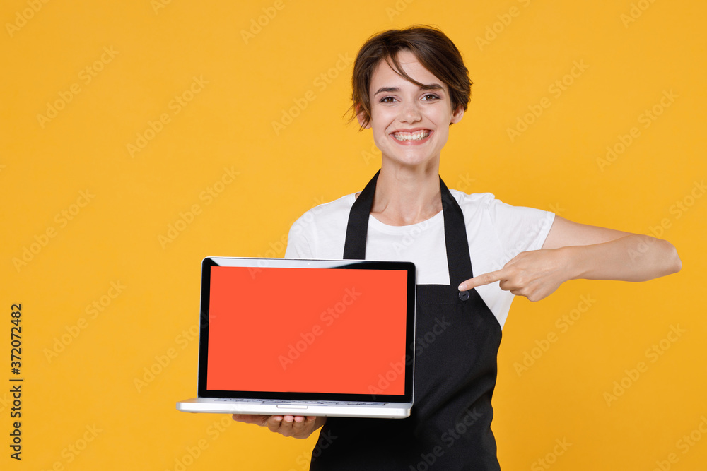 Smiling young woman 20s barista bartender barman employee in apron pointing index finger on laptop pc computer with blank empty screen mock up copy space isolated on yellow background studio portrait. Smiling young woman 20s barista bartender barman employee in apron pointing index finger on laptop pc computer with blank empty screen mock up copy space isolated on yellow background studio portrait.