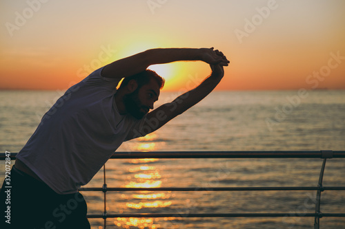 Portrait of young attractive bearded athletic man guy 20s in white t-shirt black shorts posing training doing stretching exercising warming up looking aside at sunrise over the sea outdoors.