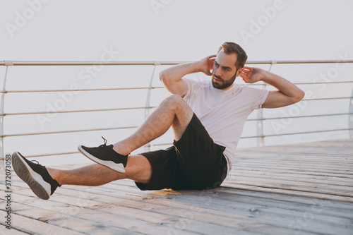 Full lenght portrait handsome attractive young bearded fitness athletic man guy 20s in white t-shirt posing training doing abdominal exercises at floor looking aside at sunrise over the sea outdoors.