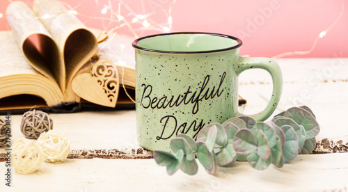 Mint tea mug. On the mug is the inscription of a beautiful day. Nearby lies an open book with heart-shaped pages. Garlands, a eucalyptus branch, decorative balls, 