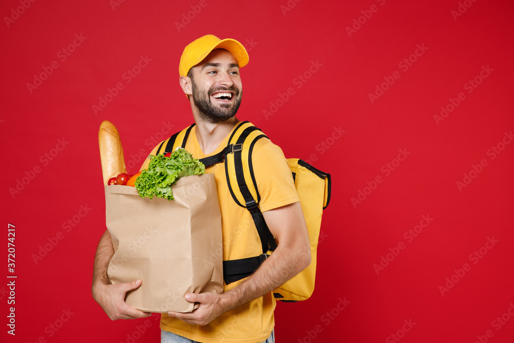 Employee courier man in yellow cap t-shirt thermal bag backpack hold brown craft paper bag takeaway with food products isolated on red background studio. Delivery service from shop, restaurant to home