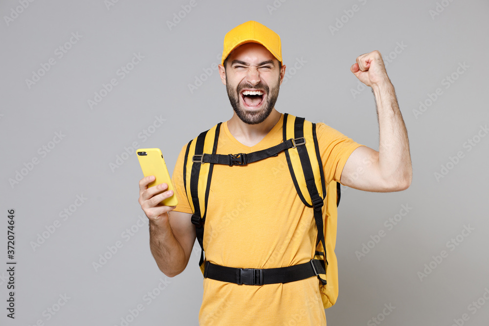 Delivery employee man guy male 20s in yellow cap t-shirt uniform thermal food bag backpack work courier service during quarantine coronavirus covid-19 hold cellphone isolated on gray background studio Delivery employee man guy male 20s in yellow cap t-shirt uniform thermal food bag backpack work courier service during quarantine coronavirus covid-19 hold cellphone isolated on gray background studio