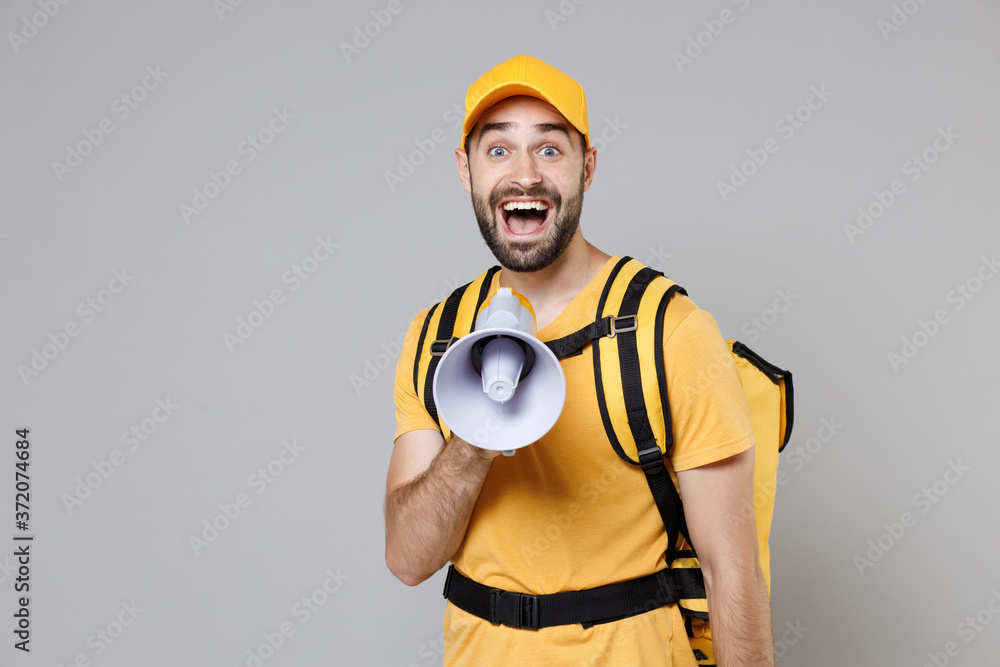 Delivery employee man guy male in yellow cap tshirt uniform thermal food bag backpack work courier service during quarantine coronavirus covid-19 scream in megaphone isolated on gray background studio Delivery employee man guy male in yellow cap tshirt uniform thermal food bag backpack work courier service during quarantine coronavirus covid-19 scream in megaphone isolated on gray background studio