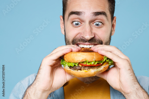 Close up of crazy young bearded man guy 20s wearing casual clothes posing holding eating american classic fast food burger looking aside isolated on pastel blue color wall background studio portrait.