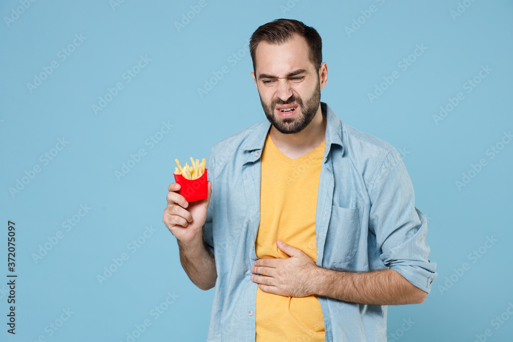 Displeased sick young bearded man guy 20s wearing casual clothes posing hold in hand french fries potatoes having ache put hand on stomach isolated on pastel blue color background studio portrait. Displeased sick young bearded man guy 20s wearing casual clothes posing hold in hand french fries potatoes having ache put hand on stomach isolated on pastel blue color background studio portrait.