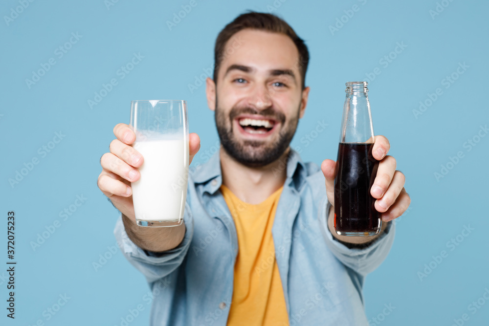 Smiling cheerful young bearded man guy 20s wearing casual clothes posing holding in hands glass of milk bottle of cola looking camera isolated on pastel blue color wall background studio portrait. Smiling cheerful young bearded man guy 20s wearing casual clothes posing holding in hands glass of milk bottle of cola looking camera isolated on pastel blue color wall background studio portrait.