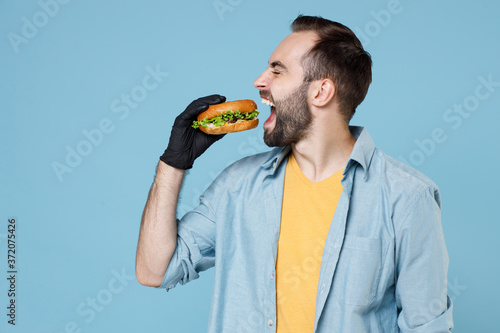 Funny young bearded man guy 20s in casual clothes black gloes posing hold biting eating american classic fast food burger keeping eyes closed isolated on pastel blue color background studio portrait.