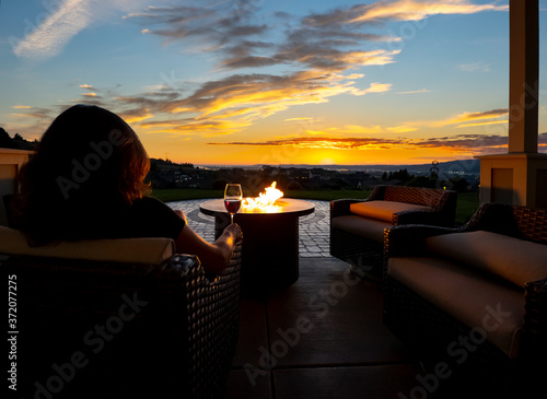 A woman relaxes with a glass of wine at night in front of an outdoor firepit on a patio of a luxury home overlooking a city and valley photo