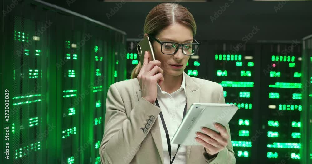 Caucasian beautiful woman among servers with tablet computer and talking on mobile phone. Pretty female analytic in data storage working on digital security, speaking on cellphone with gadget in hand. Caucasian beautiful woman among servers with tablet computer and talking on mobile phone. Pretty female analytic in data storage working on digital security, speaking on cellphone with gadget in hand.