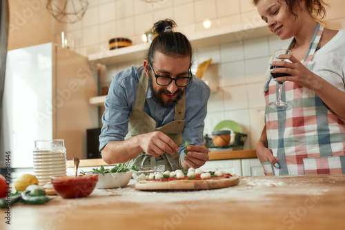 Best cook. Young couple making pizza together at home. Man in apron  professional cook adding basil on the dough while woman drinking wine. Hobby  lifestyle