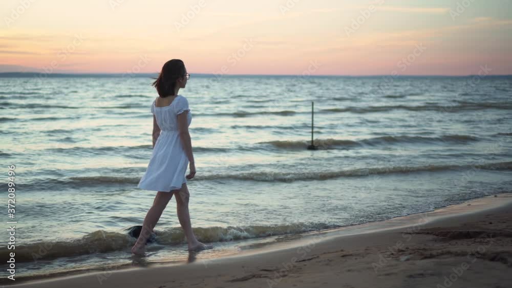 Attractive young woman walks along the sea beach at sunset. A girl in a white dress walks barefoot along the beach. Attractive young woman walks along the sea beach at sunset. A girl in a white dress walks barefoot along the beach.