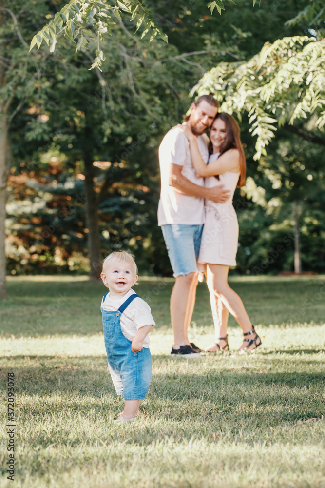 Fototapeta premium Happy smiling mother and father with baby boy in park outdoors. Family Caucasian mom and dad with son walking hugging in park on summer day. Happy family authentic lifestyle concept.