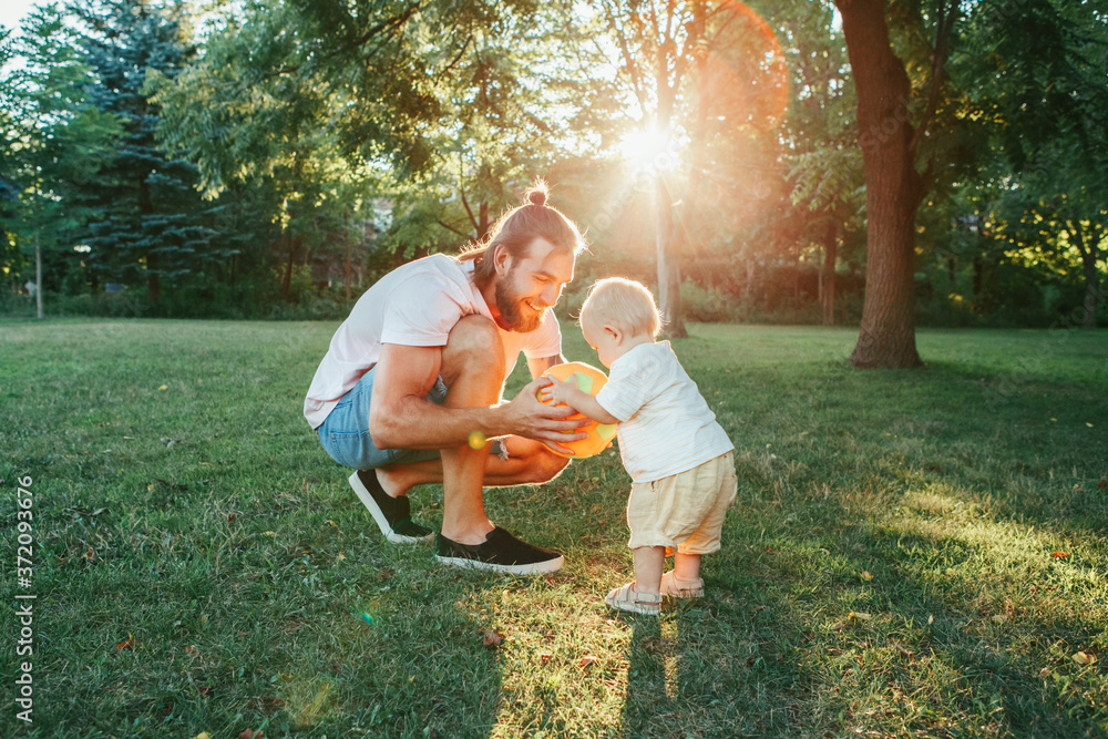 © anoushkatoronto - Young father playing ball with toddler baby boy outdoors. Parent spending time together with child son in park. Authentic lifestyle tender moment. Happy dad and active family life.
