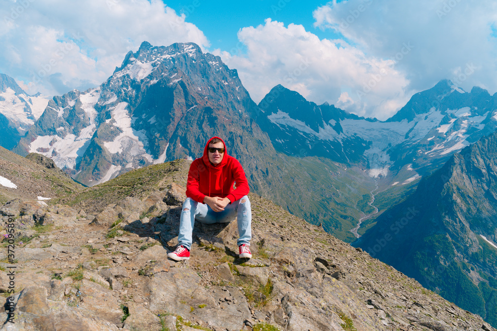 Young man in sunglasses sitting on mountain in sunny weather. Adult male in red hoodie with hood enjoying beautiful view in mountainous area.