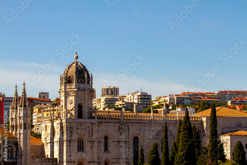 Cityscape of Lisbon, Portugal as viewed from the banks of Tagus river. Image features rooftops and exteriors of multiple buildings including the world heritage site Jeronimos Monastery.