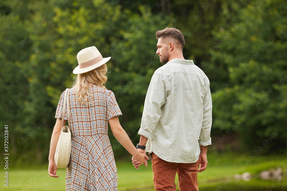 Back view portrait of romantic adult couple holding hands while walking towards river in rustic countryside scenery, copy space
