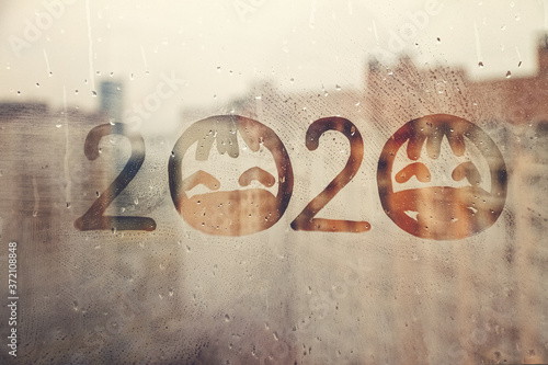 hahdmade painted numbers 2020 with sad face with closed eyes in protective medical mask on splashed by rain foggy glass on blue window, concept photo self-isolation, coronavirus, covid - 19 photo